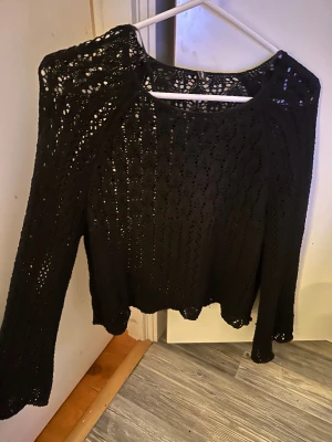 Svart stickad blus med hålmönster - Säljer en svart stickad blus med långärmad design och snyggt hålmönster. Blusen har en rund halsringning och vågig kant nedtill och vid ärmslut. Perfekt för dig som gillar en chill och trendig look med lite boho-vibe.