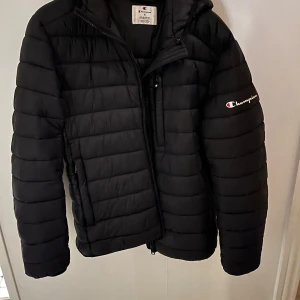 Svart pufferjacka från Champion S - Säljer en svart pufferjacka från Champion i storlek S. Jackan har horisontella quiltade paneler, huva och dragkedja framtill. Champion-logga broderad på ärmen ger en clean och sportig vibe. Perfekt för kalla dagar och enkel att matcha med allt.