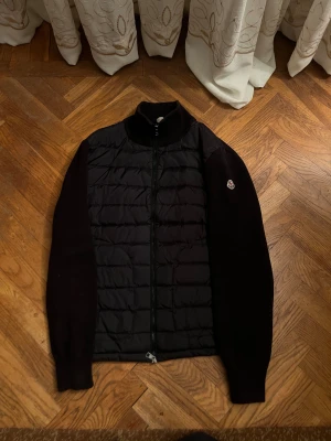 Moncler cardigan (svart) - Svart cardigan från Moncler med quiltad front och stickade ärmar. Cardiganen har hög krage, hel dragkedja och Moncler-logga på ärmen. Perfekt för dig som gillar stilren design.