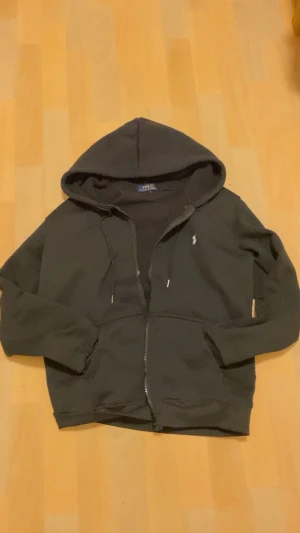 Svart hoodie från Polo Ralph Lauren - Svart hoodie från Polo Ralph Lauren med klassisk huva och snörning. Tröjan har dragkedja framtill, två fickor och den ikoniska lilla logon broderad på bröstet. Perfekt för chill dagar och enkel att matcha med jeans eller joggers.
