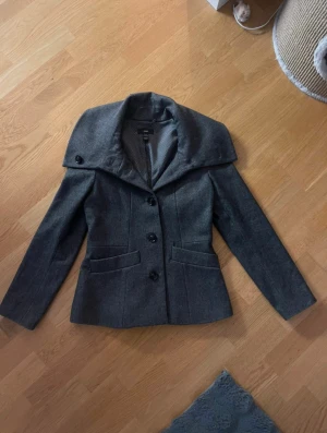 Grå kappa med krage från HM - Snygg grå kavaj från HM med bred krage och tre svarta knappar framtill. Jackan har två fickor och är figursydd för en stilren look. Perfekt för dig som gillar klassisk stil med en modern twist.