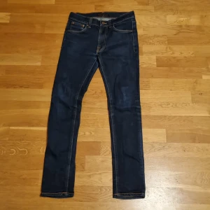 Mörkblå Nudie Jeans, storlek 29/32 - Snygga mörkblå jeans från Nudie Jeans med klassisk femficksdesign och kontrastsömmar. Modellen har raka ben och normal passform, med coola broderade detaljer på bakfickorna. Perfekt för dig som gillar stilrena och tidlösa jeans.