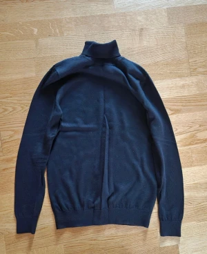 Svart polotröja John Henric M - Svart polotröja från John Henric i storlek M. Tröjan har lång ärm, ribbade muddar och klassisk hög krage. Materialet är mjukt och stickat, perfekt för kyliga dagar. Enkel och stilren design som funkar till många olika looks.