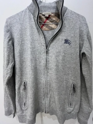 Grå cardigan från Burberry - Snygg grå cardigan från Burberry med klassisk ribbad krage och dragkedja framtill. Jackan har två fickor med dragkedja, broderad Burberry-logga på bröstet och ikoniskt rutigt foder vid halsen. Perfekt för dig som gillar stilrena och bekväma plagg.