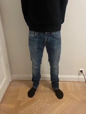 Blå jeans från Nudie med straight fit - Säljer ett par klassiska blå jeans från Nudie med snygga slitningar och broderade detaljer på bakfickorna. Modellen har straight fit, normal passning och är tillverkad i slitstarkt bomullsmaterial. Perfekt för dig som gillar en tidlös och avslappnad stil. 