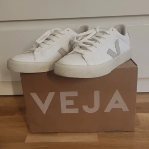 Vita Veja sneakers med grå detalj - Snygga vita sneakers från Veja med klassisk låg profil och beige V-logga på sidan. Skorna har snörning, platt sula och är tillverkade i skinn med textildetaljer. Perfekta för dig som gillar stilrena och minimalistiska sneakers.