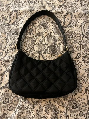 Svart quiltad axelväska med dragkedja - Snygg svart axelväska med quiltat mönster och guldfärgade detaljer. Väskan har en bred axelrem och stängs med dragkedja. Insidan har ett fack för småsaker. Perfekt storlek för det viktigaste och ger en cool vibe till din outfit.