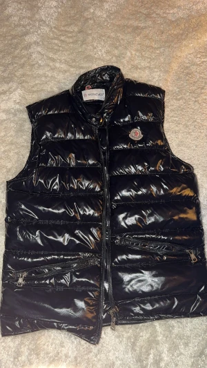 Svart dunväst från Moncler - Snygg svart dunväst från Moncler med glansig finish och klassisk logga på bröstet. Västen har hög krage, två dragkedjefickor framtill och stängs med dragkedja. Perfekt för lager-på-lager och ger en clean streetstyle-vibe.