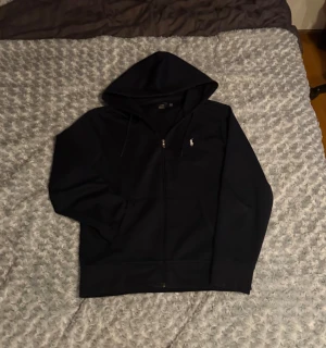 Mörkblå hoodie från Polo Ralph Lauren - Mörkblå hoodie från Polo Ralph Lauren med klassisk broderad logga på bröstet. Tröjan har huva med snörning, hel dragkedja och två fickor framtill. Materialet är mjuk bomull och passformen är normal. Perfekt för chill dagar och snygg streetstil.