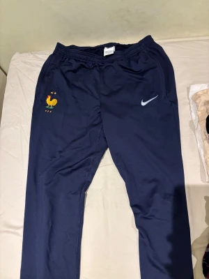 Nike Frankrike träningsbyxor blå - Mörkblå träningsbyxor från Nike med Frankrikes landslagsmärke (FFF) broderat på benet. Byxorna har dragkedjeförsedda fickor, elastisk midja och är tillverkade i ett lätt, syntetiskt material. Perfekta för fotboll eller chill.
