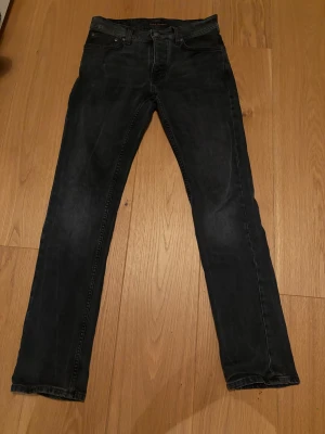 Svarta Nudie Jeans grim tim  - Säljer ett par svarta jeans från Nudie Jeans med slim fit och klassisk femficksdesign. Jeansen har knappgylf och subtila slitningar för en cool look. Materialet är mjukt denim i bomull och passformen är tight hela vägen ner.