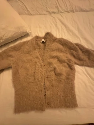 Beige fluffig kofta Holly & Whyte -  Supermjuk och fluffig beige kofta från Holly & Whyte by Lindex. Koftan har lång ärm, v-ringning och knappar framtill. Den  är tillverkad i ett pälsliknande syntetmaterial som ger en mysig känsla. 