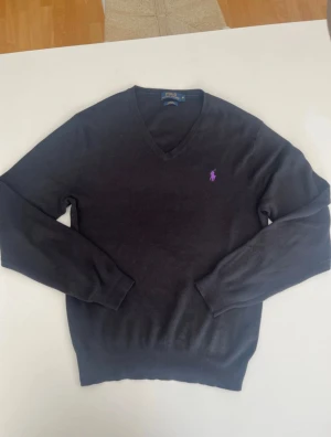 Svart v-ringad tröja Polo Ralph Lauren - Svart v-ringad tröja från Polo Ralph Lauren i slim fit med lila broderad logga på bröstet. Tillverkad i mjuk pima bomull, har långa ärmar och ribbade muddar vid ärmslut och nederkant. Perfekt för dig som gillar stilrena och klassiska plagg.