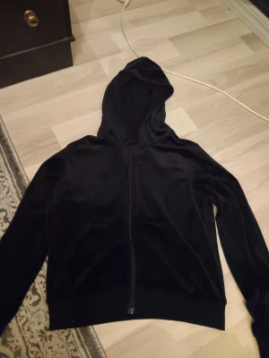 Svart hoodie i sammet med dragkedja - Svart hoodie i mjuk sammet med hel dragkedja framtill och huva. Tröjan har långa ärmar och ribbade muddar vid nederkant och ärmslut. 