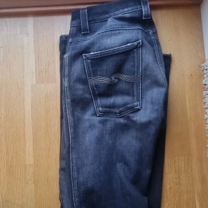 Nudie Jeans grå jeans 31/34 - Snygga blå jeans från Nudie Jeans med klassisk femficksdesign och coola slitningar framtill. Byxorna har kontrastsömmar och Nudie-loggan på bakfickan. Perfekt passform och skön känsla i mjukt denimtyg. Modell: Slim