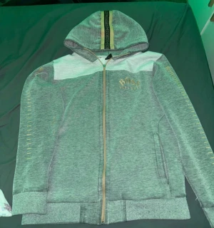 Grå hoodie från BOSS med huva S - Snygg grå hoodie från BOSS med gulddetaljer och logga på bröstet. Huva med BOSS-band i svart och guld, hel dragkedja framtill och fickor på sidorna. Långärmad och med ribbade muddar. Perfekt för chill eller streetwear.