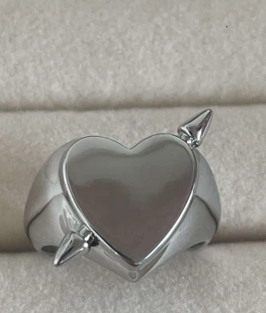 Silverring med hjärta och spjut - Cool och chunky silverring med ett blankt hjärta i mitten och två spikes på sidorna. Perfekt statement piece för dig som gillar edgy och unik stil. Ringen har en bred design och glänsande finish som drar blickarna till sig. Rostfri justerbar