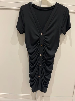 Svart ribbad kortklänning med knappar - Svart kortklänning med ribbad struktur och snygg rynkning fram och bak . Klänningen har v-ringning, korta ärmar och dekorativa knappar längs mitten. Materialet är mjukt och stretchigt,