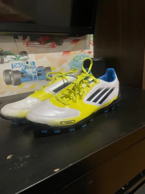 Adidas fotbollsskor vit/gul strl 43⅓ - Säljer ett par Adidas fotbollsskor i vit och gul med svarta ränder och blå detaljer på dobbarna. Skorna har snörning och är tillverkade i syntetmaterial med en låg profil och rund tå. Insidan är blå och yttersulan har rejäla dobbar för bra grepp på planen.
