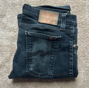 Nudie jeans - Säljer ett par svarta jeans från Nudie Jeans med klassisk design och snygga detaljer på bakfickorna. Storleken på jeansen är W30 L32. Jeansen har en slim passform och är tillverkade i mjukt denimtyg med lätt tvättad utseende.