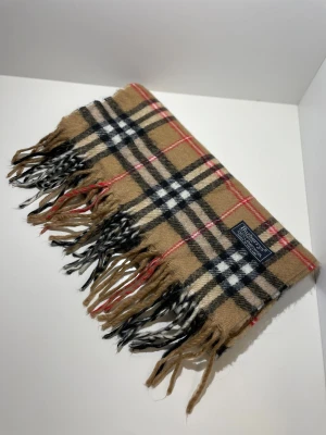 Rutig halsduk från Burberry i ull - Klassisk halsduk från Burberry i mjuk ull med det ikoniska rutiga mönstret i beige, svart, vitt och röda detaljer. Halsduken har fransar i ändarna och en snygg vävd logotypetikett. Perfekt accessoar för att lyfta din outfit.