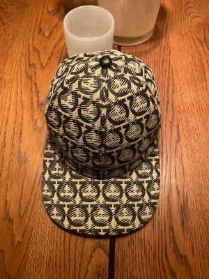 Ferragamo keps  - Snygg keps från Ferragamo med beige och svart grafiskt mönster. Kepsen har en böjd skärm, svart knapp på toppen och justerbar rem bak i svart skinn med Ferragamo-logga. Tillverkad i Italien, oanvänd. Pris kan diskuteras 