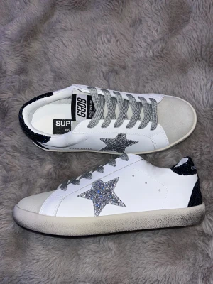 Golden Goose sneakers med glitterstjärna - Snygga vita Golden Goose sneakers med grå snörning, silverglittrig stjärna på sidan och svartglittrig häl. Ovandel i skinn och mocka, klassisk rund tå och platt sula. Perfekta för dig som vill sticka ut med lite extra bling i vardagen.