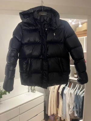 Mörkblå pufferjacka från Polo Ralph Lauren - Säljer en mörkblå pufferjacka från Polo Ralph Lauren med huva och broderad logga på bröstet. Jackan har långa ärmar med elastiska muddar, två fickor med dragkedja och quiltad design. Perfekt för kalla dagar och riktigt snygg stil. Barnstorlek XL ca 155-160cm. 