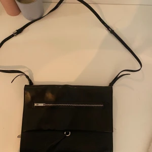 Svart axelväska från Tiger of Sweden - Snygg svart axelväska från Tiger of Sweden i skinn. Väskan har justerbar axelrem, en dragkedja framtill och stilren design med både släta och strukturerade partier. Perfekt för dig som vill ha en klassisk och tidlös look.