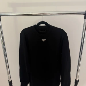 Svart sweatshirt från Prada - Stilren svart sweatshirt från Prada med klassisk rund halsringning och diskret logga i metall framtill. Tröjan har långa ärmar och en enkel, clean look som passar perfekt till streetwear eller en mer dressad stil.