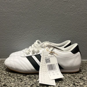 Adidas Taekwondo vita sneakers - Snygga Adidas Taekwondo sneakers i vitt skinn med klassiska svarta ränder på sidorna och gummisula. Skorna har snörning och låg profil, perfekt för dig som gillar sportig och clean stil. Adidas-logga på plösen och insidan.