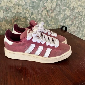 Adidas Campus rosa sneakers mocka - Säljer ett par Adidas Campus sneakers i rosa mocka med vita detaljer och klassiska tre ränder på sidorna. Skorna har vit sula, breda vita skosnören och Adidas logga på häl och plös. Perfekta för dig som gillar retrostil och vill sticka ut lite extra.