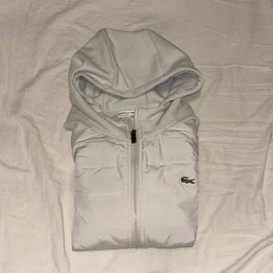 Ljusgrå hoodie från Lacoste med dragkedja - Säljer en ljusgrå hoodie från Lacoste med dragkedja framtill och två fickor. Tröjan har quiltad framsida och baksida, klassisk huva och den ikoniska krokodilloggan på bröstet. Perfekt för kyliga dagar och snygg till jeans eller joggers.