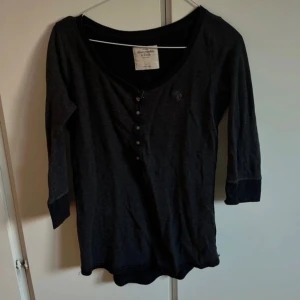 Svart topp från Abercrombie & Fitch - Svart topp från Abercrombie & Fitch med trekvartsärm och rund halsringning. Toppen har knappar framtill och en liten broderad logga på bröstet. Tillverkad i mjukt bomullsmaterial, perfekt för en chill och stilren look.