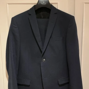 Mörkblå ull-kostym - Tiger of Sweden - Säljer en stilren mörkblå kostym från Tiger of Sweden, modell JILE. Kavajen har klassisk passform med två fickor och enkla knappar. Byxorna har pressveck och bakficka med knapp. Materialmix av ull, polyester, polyamid och elastan för en snygg look. (Köparen står för frakten)