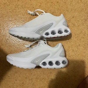 Nike Air Max DN vita sneakers - Nike Air Max DN sneakers i helvitt med mesh-överdel och futuristisk design. Snygga detaljer i silver och transparenta Air Max-enheter i sulan. Skorna har snörning och en platt, greppvänlig sula med röda detaljer undertill. Perfekta för dig som gillar modern streetstyle. Aldrig använda pris kan diskuteras vid snabb affär.