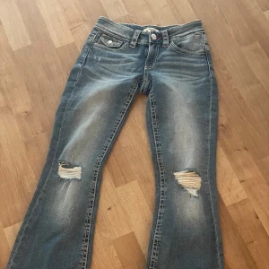 Blå bootcut jeans med slitningar - Snygga blå jeans med bootcut passform och slitna detaljer på knäna. Jeansen har klassisk femficksdesign, röda broderier på bakfickorna och en cool tvättad look. Perfekta för dig som gillar en avslappnad och trendig stil. Aldrig använt!!!