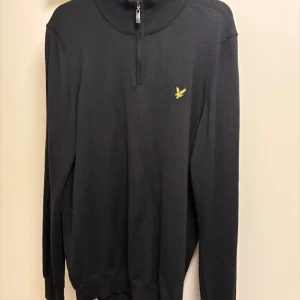 Svart half zip tröja från Lyle & Scott - Svart långärmad tröja från Lyle & Scott med half zip och gul broderad logga på bröstet. Tröjan har ribbad krage och är tillverkad i mjukt stickat material. Perfekt för en stilren och avslappnad look.