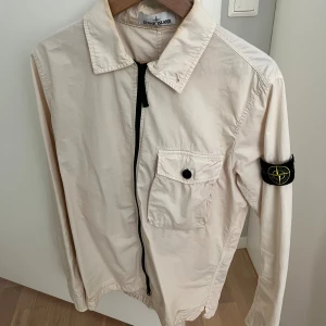 Beige overshirt från Stone Island - Säljer en beige overshirt från Stone Island med klassisk krage, bröstficka med knapp och ikonisk patch på ärmen. Jackan har dragkedja framtill och är tillverkad i bomull. Perfekt för lager-på-lager och clean streetstyle. Jackan är strl M, i 8/10 skick och varsamt använd. Säljer för 1000kr, men kan gå ner i snabb affär. 