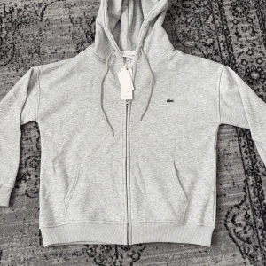 Grå huvud tröja från Lacoste - Snygg ljusgrå hoodie från Lacoste med klassisk krokodillogga på bröstet. Tröjan har huva med snörning, två fickor framtill och hel dragkedja. Tillverkad i mjuk bomullsmix och har ribbade muddar vid ärmslut och nederkant.