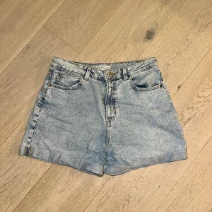 Högmidjade ljusblå jeansshorts - Säljer ett par klassiska högmidjade jeansshorts i ljusblå denim. De har fem fickor, uppvikta benslut och bälteshällor. Perfekta för stranden eller sommardagar. Snygg vintagekänsla och medium loose fit som ger en avslappnad vibe.