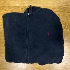 Mörkblå hoodie från Polo Ralph Lauren - Mörkblå hoodie med dragkedja från Polo Ralph Lauren. Klassisk design med huva och broderad röda logga på bröstet. Tillverkad i mjuk bomull, perfekt för chill dagar eller när du vill ha en clean streetstyle.