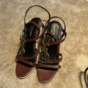 Bruna sandaler med kilklack - Snygga bruna sandaler med ikoniskt emblem i metall framtill. Skorna har flera smala remmar i skinn och en hög kilklack i repmaterial. Perfekta för att lyfta din sommarstil med en lyxig touch.