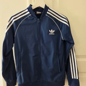 Blå Adidas zip-tröja - Säljer en klassisk zip-tröjq från Adidas med vita ränder längs ärmarna och logga på bröstet. Tröjan har dragkedja framtill, ribbade muddar och två fickor. Perfekt för dig som gillar sportig stil och streetwear.