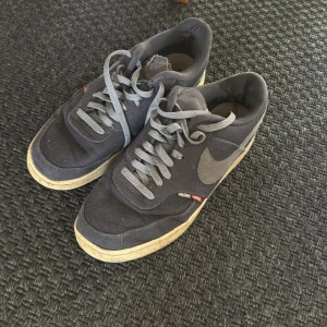 Nike Air Force mörkgrå sneakers - Mörkgrå Nike Air Force sneakers med klassisk siluett och grå swoosh på sidan. Skorna har snörning och platt sula, samt detaljer i textil och syntet. Perfekta för dig som gillar streetstyle och vill ha en ikonisk sneaker i garderoben.