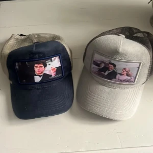 Två truckerkepsar med filmtryck - Säljer två coola truckerkepsar från Muggiori med nät baktill. Ena kepsen är mörkblå med ett filmtryck på framsidan, den andra är ljusgrå med ett annat filmtryck. Perfekta accessoarer för dig som gillar unika detaljer och filmreferenser.