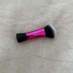 Real Techniques mini sculpting brush - En mini medium sculpting brush från Real Techniques med ett metallicrosa handtag och svart gummigrepp. Borsten är tät, snedskuren och har svart-vita syntetiska strån, perfekt för contouring och bronzer.