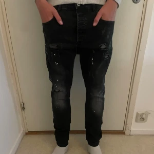 Svarta skinny jeans från Kings Will Dream - Svarta jeans med skinny fit och coola slitningar samt vita färgstänk på framsidan. Jeansen har klassisk femficksdesign och knappgylf. Materialet är stretchigt denim som sitter tajt längs benen och ger en modern look. Storlek 32/31