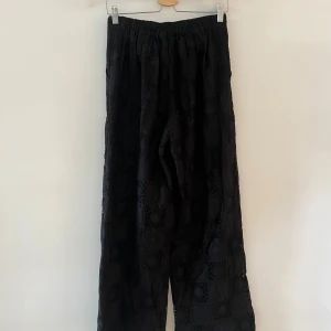 Svarta vida spetsbyxor med mönster - Svarta byxor med bred passform och snyggt spetsmönster i hela tyget. Byxorna har resår i midjan och ett detaljerat, rutigt spetsmönster som ger en cool och unik look. Perfekta för dig som vill sticka ut med en trendig och bekväm stil. Skicka ett meddelande för fler bilder.