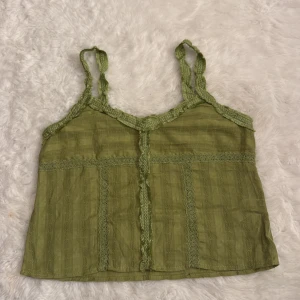 Grön linnetopp med spetsdetaljer - Säljer en ljusgrön linnetopp med tunna axelband och fina spetsdetaljer längs kanterna och framtill. Toppen har ett diskret rutigt mönster i tyget och är lätt croppad. Perfekt för varma dagar och enkel att styla med jeans eller kjol.